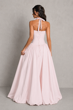 Woman wearing a long, light pink dress with a halter neck design against a plain background.