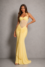 A woman modeling a pale yellow halter-neck maxi dress with a sheer mesh bodice and dramatic gathered draping on the skirt, posed against a textured gray studio backdrop.