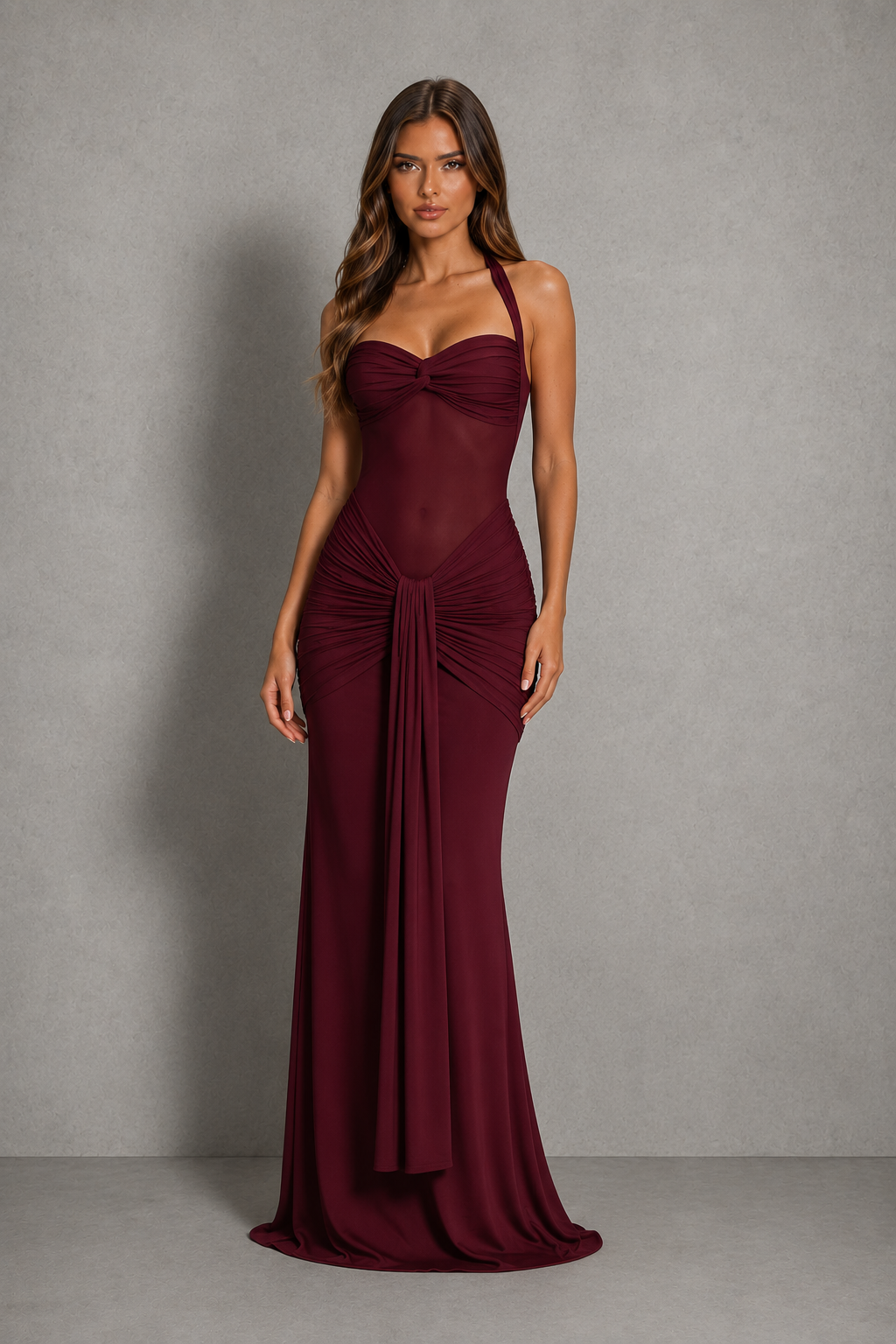 A woman modeling a floor-length burgundy gown with a sweetheart neckline, halter straps, ruched bodice, and draped skirt against a textured gray studio backdrop.