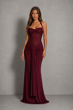 A woman modeling a floor-length burgundy gown with a sweetheart neckline, halter straps, ruched bodice, and draped skirt against a textured gray studio backdrop.