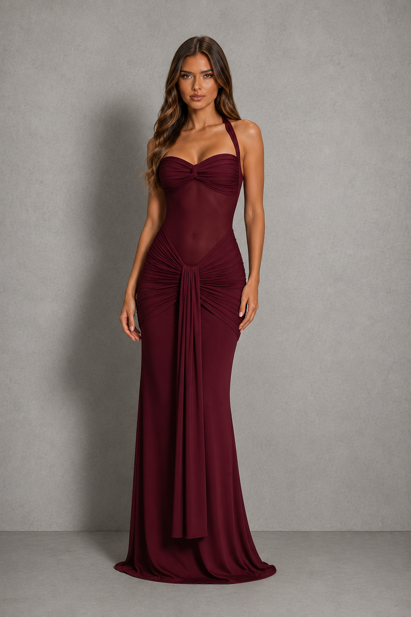 A woman modeling a floor-length burgundy gown with a sweetheart neckline, halter straps, ruched bodice, and draped skirt against a textured gray studio backdrop.