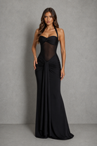 A woman modeling a floor-length black gown with a sheer mesh bodice, halter-style straps, and a gathered skirt, standing against a textured gray studio backdrop.