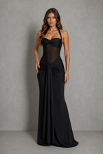 A woman modeling a floor-length black gown with a sheer mesh bodice, halter-style straps, and a gathered skirt, standing against a textured gray studio backdrop.