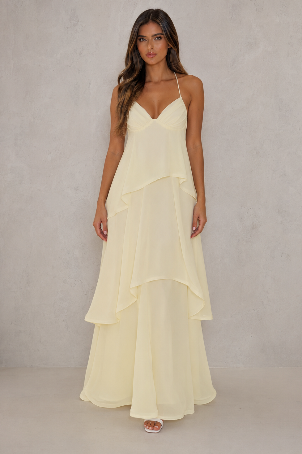 A woman modeling a cream-colored, sleeveless maxi dress with thin spaghetti straps, a V-neckline, and a layered, ruffled tiered skirt, set against a textured light gray studio backdrop.