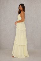 A woman modeling a pale yellow, tiered halter-neck maxi dress against a textured gray studio backdrop.