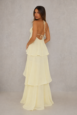 A woman modeling a pale yellow tiered maxi dress from the back, featuring thin straps, a low open back with tie details, and a flowing layered skirt, set against a textured gray studio backdrop.