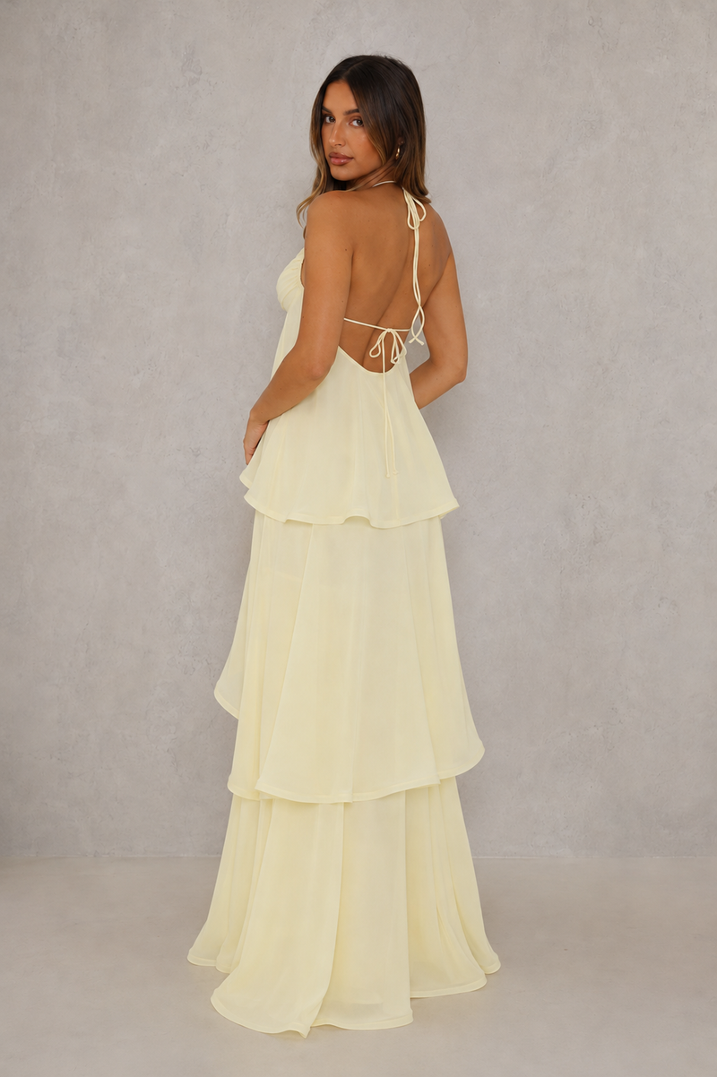 A woman modeling a pale yellow tiered maxi dress from the back, featuring thin straps, a low open back with tie details, and a flowing layered skirt, set against a textured gray studio backdrop.
