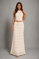 A woman modeling a two-piece cream or off-white textured maxi dress with a sleeveless high-neck crop top and a long tiered skirt, set against a plain gray studio background.