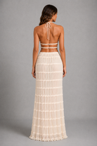 A woman shown from the back wearing a cream-colored two-piece outfit consisting of a halter crop top and a long pleated skirt, set against a solid gray studio background.