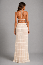 A woman shown from the back wearing a cream-colored two-piece outfit consisting of a halter crop top and a long pleated skirt, set against a solid gray studio background.