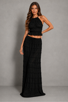 A woman modeling a black two-piece outfit consisting of a halter-neck crop top with a ruffled hem and a matching long, tiered maxi skirt, set against a plain gray studio background.