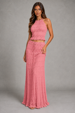 A woman modeling a two-piece pink knit set consisting of a sleeveless halter crop top and a matching high-waisted maxi skirt, set against a neutral gray studio background.