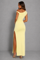 A woman modeling a pale yellow off-the-shoulder maxi dress from the back, featuring a high thigh slit and ruched detailing, set against a plain gray studio background.