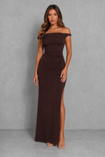 A woman modeling a dark brown off-the-shoulder maxi dress with a high side slit, standing against a textured gray studio backdrop.