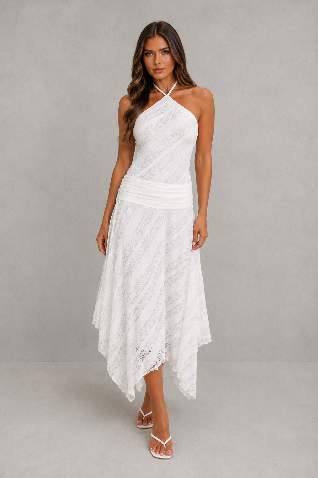 A woman modeling a white lace halter-neck midi dress with an asymmetrical hem, paired with white thong sandals, against a neutral gray studio background.