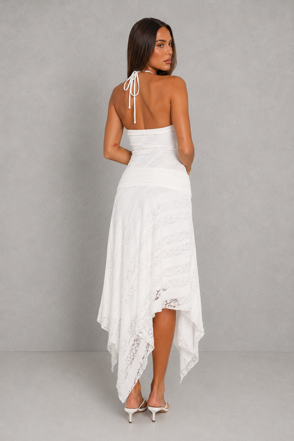 A woman modeling a white halter-neck high-low dress from the back, featuring a lace-trimmed hem and tie-back straps, set against a neutral gray studio background.