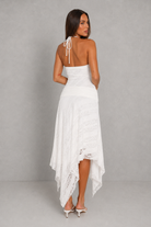 A woman modeling a white halter-neck high-low dress from the back, featuring a lace-trimmed hem and tie-back straps, set against a neutral gray studio background.