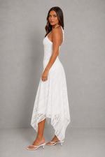 A woman modeling a white sleeveless halter-neck maxi dress with a high-low hem and lace trim, paired with white heeled sandals, against a gray studio background.