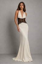 A woman modeling a floor-length halter gown with a deep V-neckline, fitted silhouette, and a contrasting black lace waistband over an ivory satin skirt, set against a neutral gray studio backdrop.