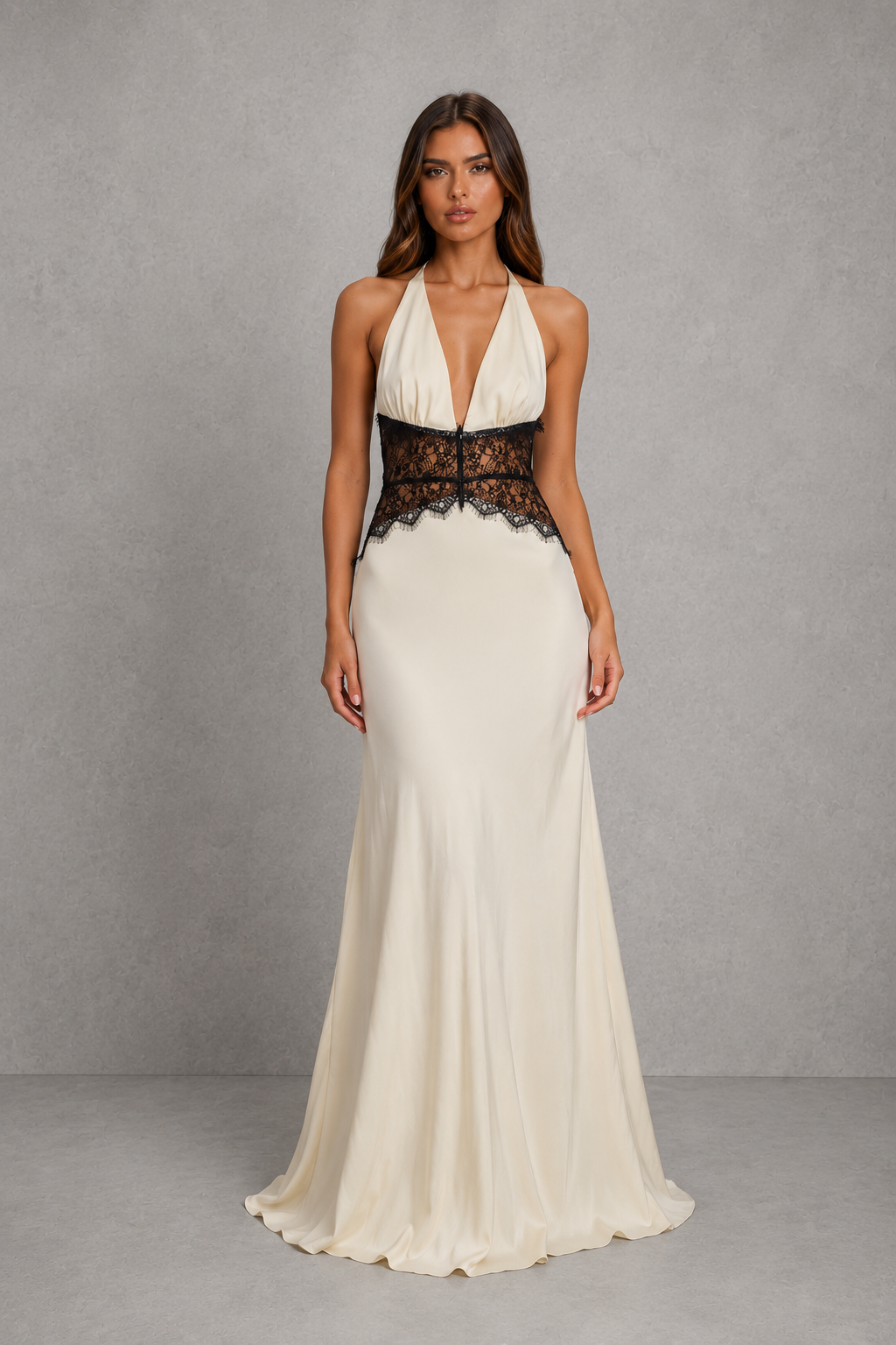 A woman modeling a floor-length cream gown with a deep V-neckline, halter-style shoulders, and a wide black lace waistband, photographed against a textured gray studio backdrop.