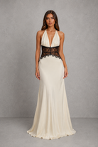A woman modeling a floor-length cream gown with a deep V-neckline, halter-style shoulders, and a wide black lace waistband, photographed against a textured gray studio backdrop.