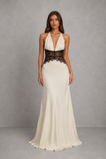 A woman modeling a floor-length cream gown with a deep V-neckline, halter-style shoulders, and a wide black lace waistband, photographed against a textured gray studio backdrop.