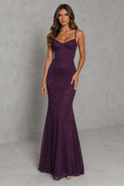 A woman modeling a sleeveless, fitted mermaid-style gown in deep plum purple lace, standing against a textured gray studio backdrop.