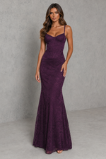 A woman modeling a sleeveless, fitted mermaid-style gown in deep plum purple lace, standing against a textured gray studio backdrop.