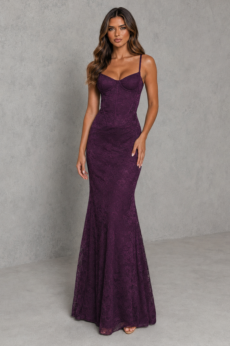 A woman modeling a sleeveless, fitted mermaid-style gown in deep plum purple lace, standing against a textured gray studio backdrop.
