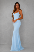 A woman modeling a light blue spaghetti-strap maxi dress with a fitted bodice and flared skirt, standing against a textured gray studio wall.