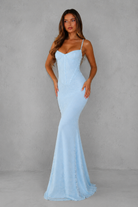 A woman modeling a light blue spaghetti-strap mermaid gown against a textured gray studio backdrop.