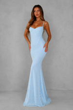 A woman modeling a light blue spaghetti-strap mermaid gown against a textured gray studio backdrop.