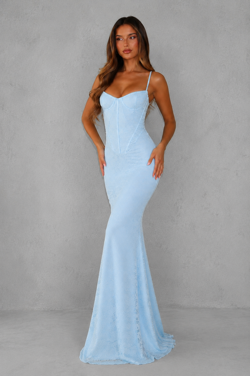 A woman modeling a light blue spaghetti-strap mermaid gown against a textured gray studio backdrop.