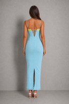 A woman viewed from the back wearing a light blue maxi dress with thin straps, a lace-up open back, and a front slit, paired with white high heels against a gray studio background.