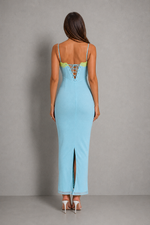 A woman viewed from the back wearing a light blue maxi dress with thin straps, a lace-up open back, and a front slit, paired with white high heels against a gray studio background.