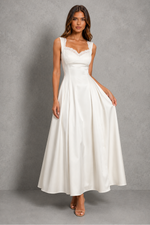 A woman modeling a sleeveless white maxi dress with a sweetheart neckline, lace trim, and a full satin skirt, standing against a textured gray studio backdrop.