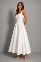 A woman modeling a sleeveless white A-line maxi dress with a V-neckline and gathered waist, standing against a textured gray studio backdrop.