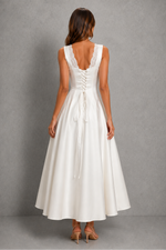 A woman viewed from the back wearing a sleeveless white maxi dress with lace-up back detailing and a flared skirt, standing against a textured gray studio backdrop.