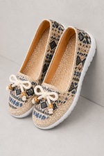 A pair of women's slip-on shoes with a beige woven snakeskin pattern, featuring white rope bows with small gold and white beads on the toes, and white soles.