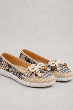 A pair of women's slip-on shoes with a beige and black snakeskin pattern, featuring white beaded bow details on the toe and white soles, set against a light gray textured background.