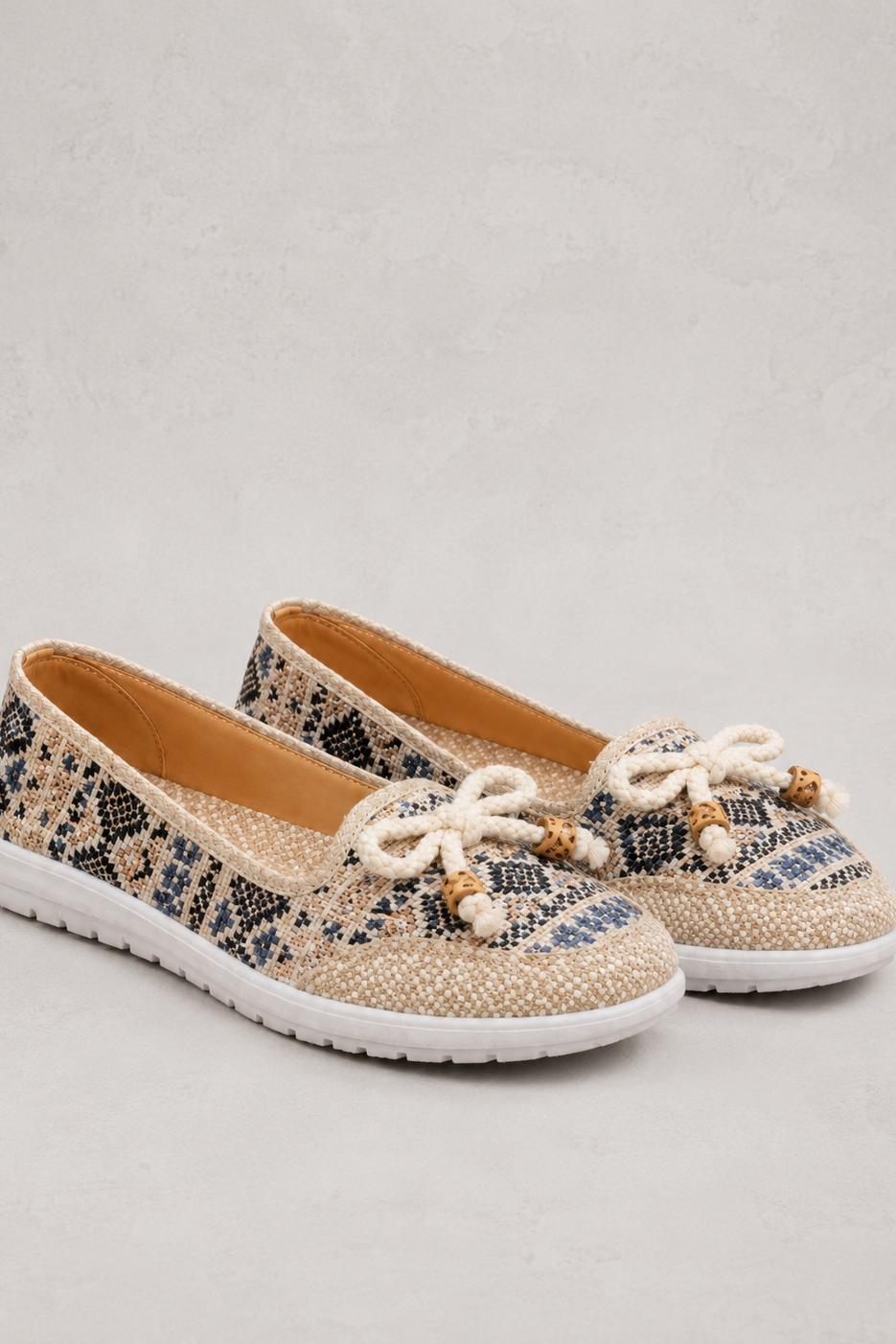 A pair of women's slip-on shoes with a beige and black snakeskin pattern, featuring white beaded bow details on the toe and white soles, set against a light gray textured background.