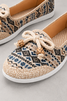 A pair of woven slip-on shoes with a geometric pattern in beige, black, and blue, featuring a thick cream rope bow with wooden beads on the front and a white sole.