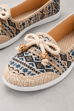 A pair of woven slip-on shoes with a geometric pattern in beige, black, and blue, featuring a thick cream rope bow with wooden beads on the front and a white sole.