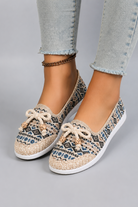 A pair of women's slip-on flat shoes with a woven geometric pattern in beige, blue, and black, featuring cream-colored bows on the toes, shown on a model wearing light-wash jeans against a gray studio background.