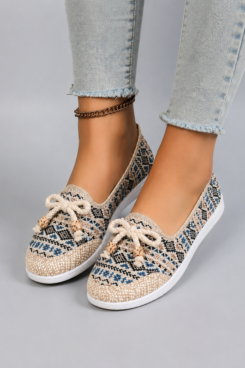 A pair of women's slip-on flat shoes with a woven geometric pattern in beige, blue, and black, featuring cream-colored bows on the toes, shown on a model wearing light-wash jeans against a gray studio background.