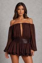A woman modeling an off-the-shoulder dark brown long-sleeve romper with a deep V-neckline, pleated texture, and a wide matching belt, set against a textured gray studio background.
