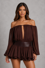 A woman modeling an off-the-shoulder dark brown long-sleeve romper with a deep V-neckline, pleated texture, and a wide matching belt, set against a textured gray studio background.