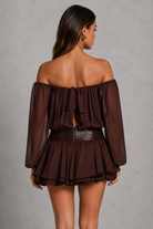 A woman shown from the back wearing a dark brown off-the-shoulder mini dress with sheer long sleeves, a ruffled tie detail at the back, and a textured waistband, set against a neutral gray studio background.