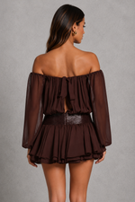 A woman shown from the back wearing a dark brown off-the-shoulder mini dress with sheer long sleeves, a ruffled tie detail at the back, and a textured waistband, set against a neutral gray studio background.