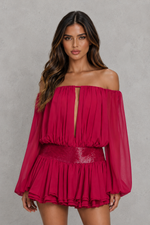 A woman modeling a bright fuchsia off-the-shoulder mini dress with long sheer sleeves, a keyhole neckline, and a ruffled peplum skirt against a textured gray studio background.